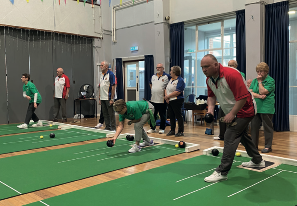 Players compete in an indoor bowls match on green mats inside a community hall. Several participants in green and red team shirts are in action, rolling bowls toward the yellow jack, while others in blue and white shirts watch from the side. The hall is decorated with bunting, and the atmosphere appears focused and friendly.