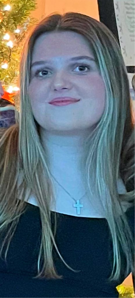 a woman with long blonde hair wearing a black top and a cross necklace, softly lit in front of a warm background.