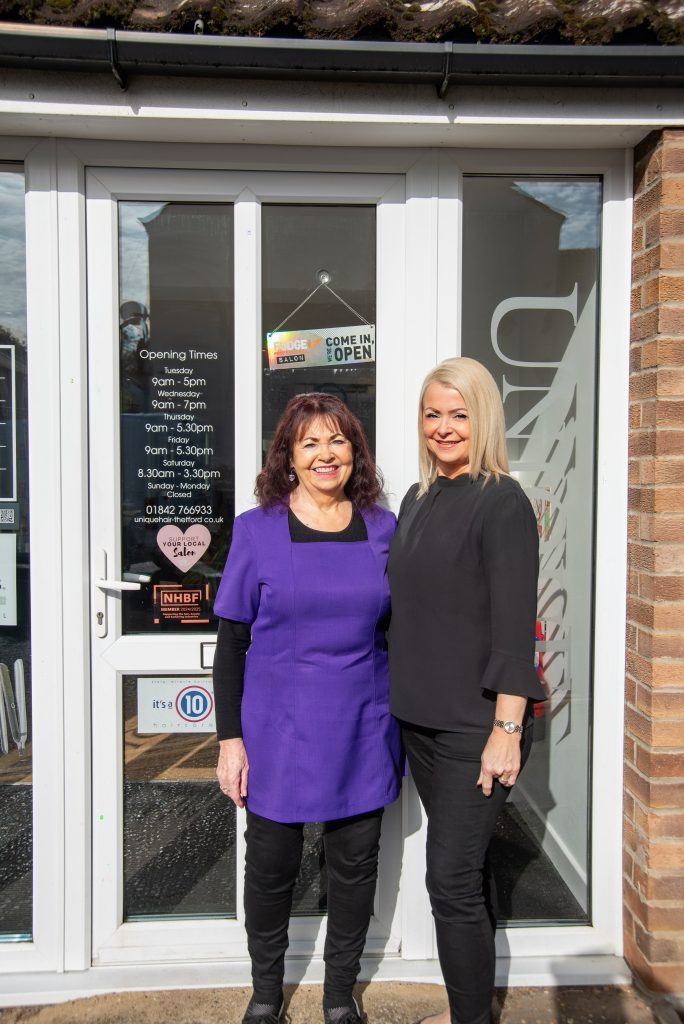 Two women outside Unique Hair & Beauty – both smiling, standing in front of the salon entrance with “Come in, we’re open” sign visible.