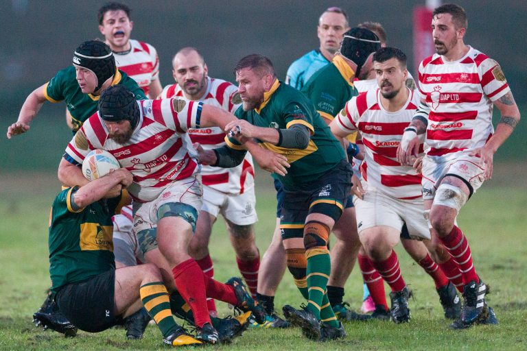 A group of rugby players in the middle of a match. Players wearing red and white striped jerseys clash with those in green and yellow jerseys as they compete for possession of the ball on a muddy field. The scene is intense and action-packed, with players pushing, tackling, and running.