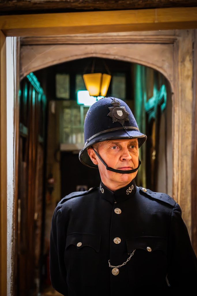 A person dressed in a traditional British police uniform, including a dark tunic and a tall helmet with a badge, stands in a narrow hallway lit by warm overhead lights, looking slightly to the side with a serious expression.