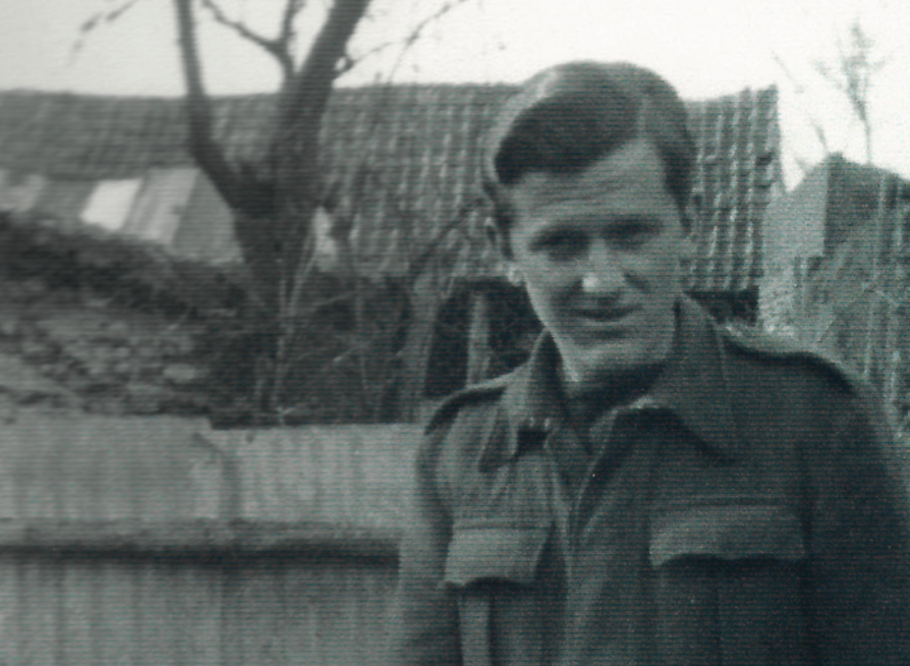 A black-and-white photograph of a young man in military uniform standing outdoors, looking toward the camera. Behind him is a simple fence and the roof of a building.