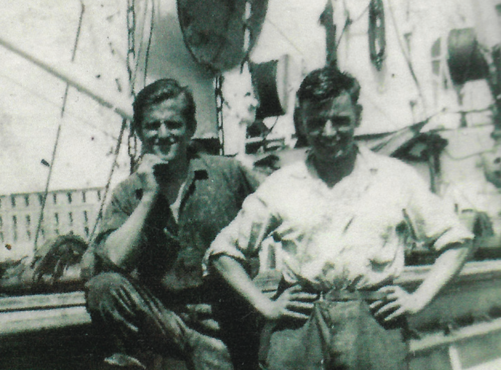 A black-and-white photo showing two young men smiling aboard a ship. Both wear rolled-up shirts and work clothes, posing casually among ropes and rigging.