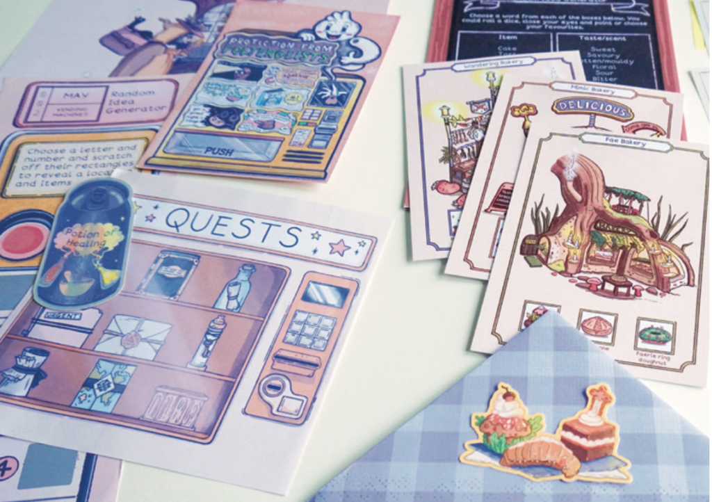 A close-up photo of a colourful tabletop game setup. Illustrated cards, sheets, and stickers are scattered across a light surface, featuring pastel designs of fantasy-style shops, potions, and quest icons. One card reads “Protection from Problem Beasts,” while another shows a section labelled “Quests.” A small sticker of pastries and a croissant decorates a blue gingham-patterned envelope in the corner. The overall aesthetic is whimsical and hand-drawn, with a focus on creative, fantasy-inspired artwork.