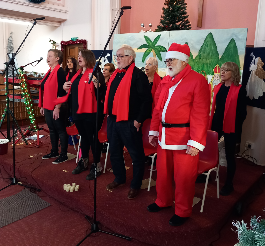A festive indoor performance scene showing a mixed-age choir standing in a church hall, singing into microphones on stands. The singers are dressed mainly in black with bright red scarves, forming a line across the front of the room. One older man on the right is dressed as Father Christmas, wearing a red Santa suit, white beard, glasses and hat. Behind the choir is a painted nativity-style backdrop with palm trees, hills and figures, alongside a decorated Christmas tree. The space features red carpeting, chairs behind the performers, microphone cables on the floor, and Christmas decorations including tinsel and lights, creating a warm, seasonal community concert atmosphere.