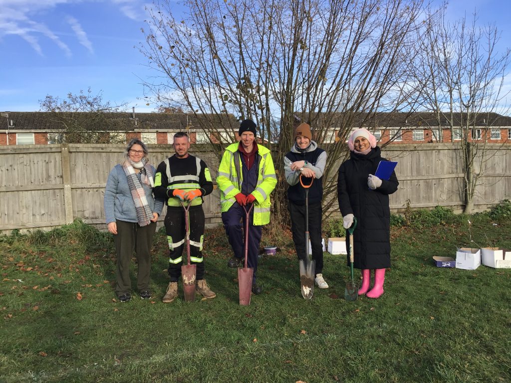 A group of six adults standing outdoors on grass in winter daylight, posing in a line and holding spades. They are dressed in warm clothing, including coats, hats, gloves, and workwear, suggesting a cold day. One person wears a high-visibility jacket, and another wears bright pink wellington boots. Behind them is a wooden fence, leafless trees, and a row of brick houses. Cardboard boxes and planting supplies are visible on the ground to the right, indicating a community planting or outdoor volunteering activity.