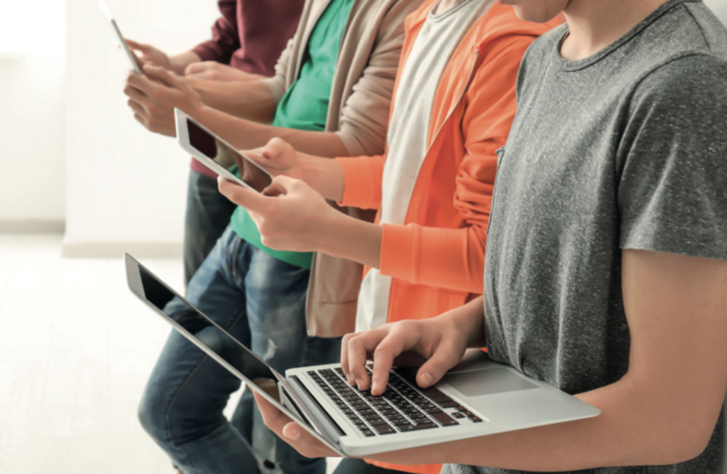 A stock-style photograph showing a row of young people standing side by side indoors, each using a different digital device. In the foreground, one person holds an open laptop and types on the keyboard, while others beside them hold smartphones and a tablet, looking down at their screens. Only torsos and hands are visible, with casual clothing including T-shirts, hoodies, and jeans in muted colours. The background is plain and softly lit, keeping the focus on the devices and the act of using technology.