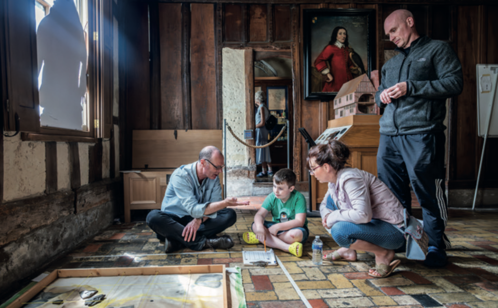 A magazine-style promotional image showing an activity taking place inside a historic timber-framed building. The scene is set in a wood-panelled room with exposed beams, leaded windows and a patterned tiled floor. In the centre, a man sits cross-legged on the floor, leaning forward and gesturing as he explains something to a young child seated opposite him. The child sits on the floor with a pencil and worksheet, listening intently. Two adults crouch nearby, watching and engaging with the activity, while another adult stands slightly behind them holding a folded leaflet or model. On the floor in the foreground is a large framed board with strings and markings laid out, suggesting a hands-on learning or investigative activity. In the background, a doorway leads to another room where a visitor can be seen, and a framed historical portrait hangs on the wall above a wooden display stand, reinforcing the heritage setting. Natural light streams in from a window on the left.