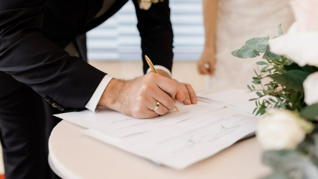 A close-up photograph showing a formal document being signed on a table. In the foreground, a person wearing a dark suit jacket signs paperwork with a gold pen, their hand and wedding ring clearly visible. A second person stands just behind, partially visible and dressed in light-coloured formal clothing. A small floral arrangement with white flowers and green foliage sits on the right-hand side of the table. The background is softly blurred, keeping the focus on the act of signing the document.