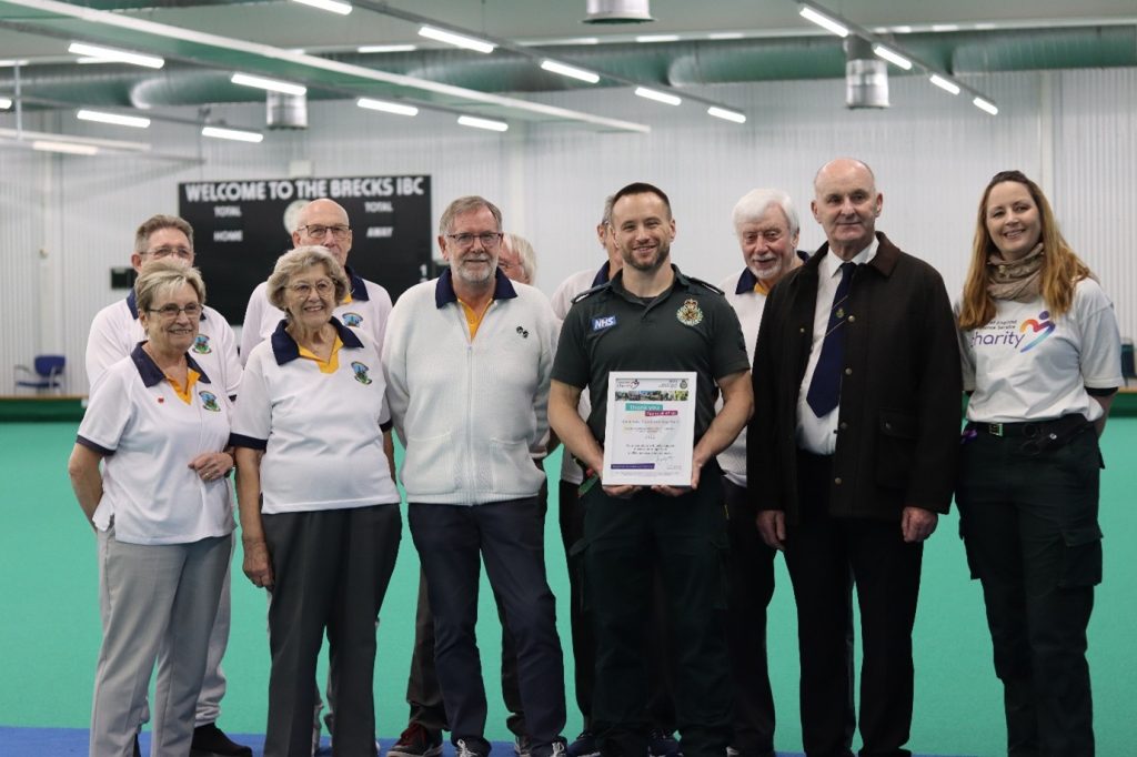 A group photograph taken indoors at Breckland Indoor Bowls Club, with a sign reading “Welcome to the Brecks IBC” visible in the background. A group of adults stand together on the green, including several people wearing matching white polo shirts with a bowls club logo. At the centre, a man in a green NHS uniform holds a certificate. Other people in the group are wearing formal jackets, ties, or charity-branded clothing. The image shows the group smiling and posing for the camera inside the sports hall, with overhead lighting and the bowling surface clearly visible.