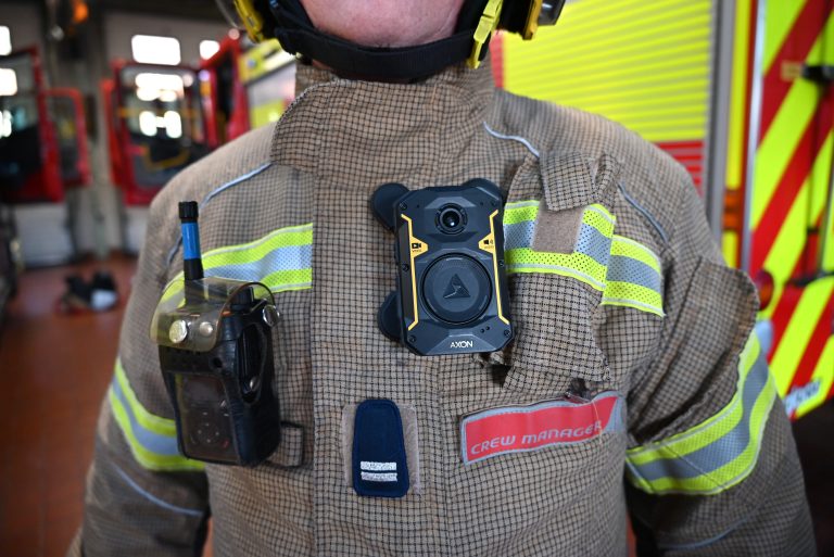 Close-up of a firefighter wearing protective turnout gear inside a fire station. A black Axon body-worn camera is mounted on the front of the jacket, alongside a radio clipped to the chest. Reflective yellow safety stripes run across the coat, and a red name badge reads “Crew Manager.” Fire engines with high-visibility markings are visible blurred in the background.