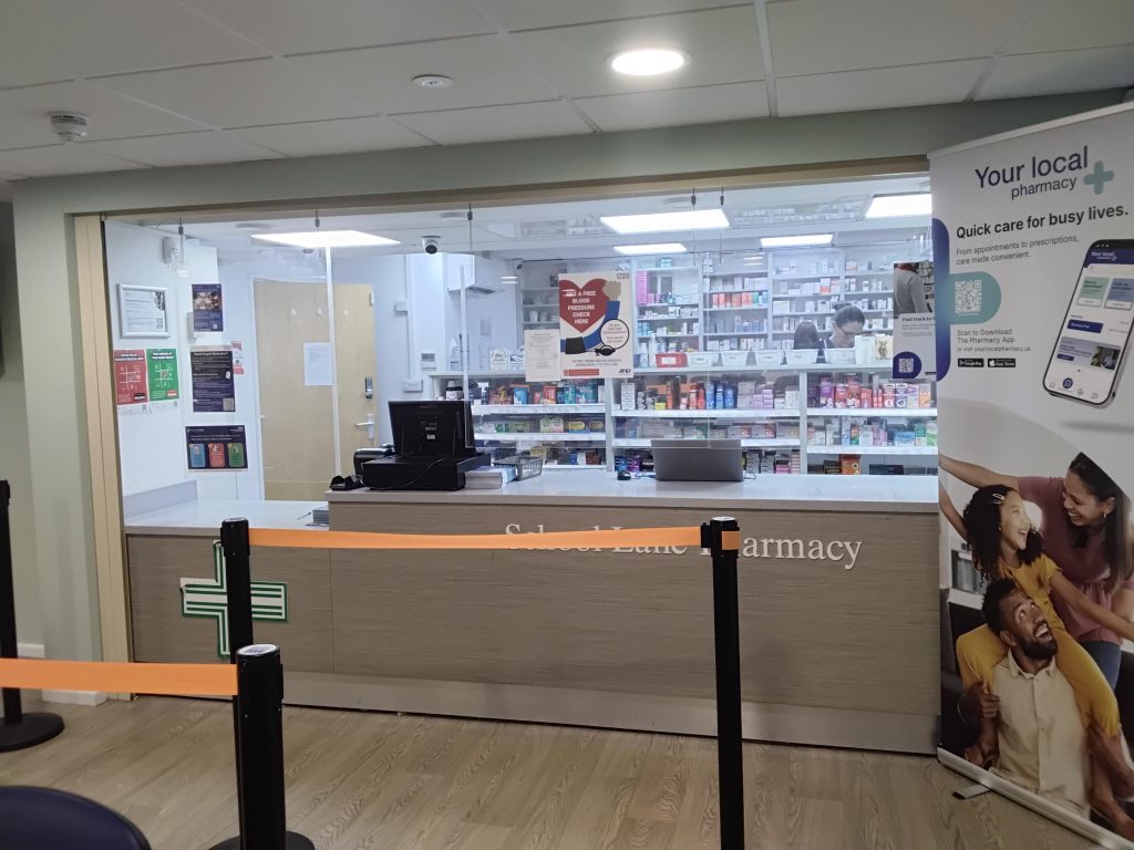 Interior view of a local pharmacy counter area. A reception desk with a green pharmacy cross symbol faces the entrance, with black queue posts and orange barrier tape in front. Behind a glass screen, shelves are stocked with medicines and healthcare products, and two staff members are visible working. Posters and notices are displayed on the walls, and a promotional banner to the right advertises a pharmacy app and quick care services.
