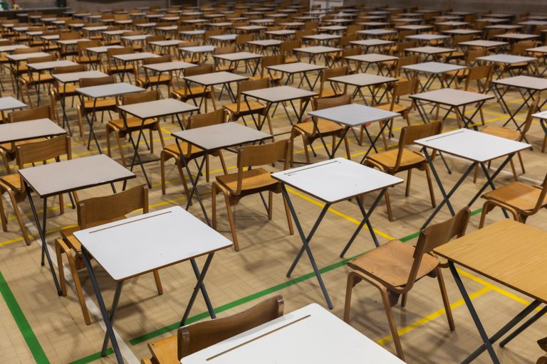 A large exam hall filled with rows of individual desks and wooden chairs spaced evenly apart. Each desk has a light-coloured writing surface and metal legs, arranged in a grid pattern across a marked sports hall floor with yellow and green lines. The room is empty, prepared for students to sit exams.