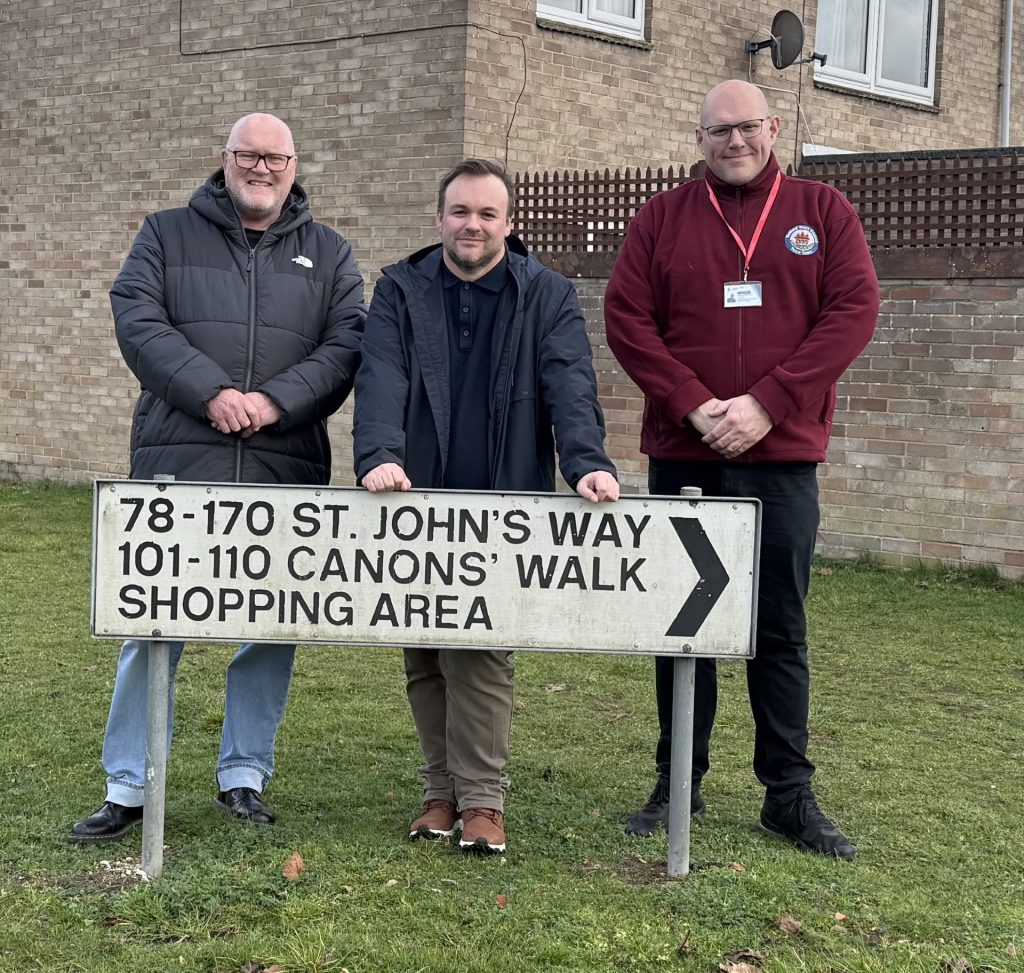 Three men stand on grass in front of a brick residential building, posing behind a white road sign that reads: “78–170 St. John’s Way, 101–110 Canons Walk, Shopping Area” with a right-pointing arrow. The man on the left wears glasses and a dark padded jacket; the man in the centre wears a dark coat and rests his hands on the top of the sign; the man on the right wears a burgundy zip-up top with an ID badge on a red lanyard.