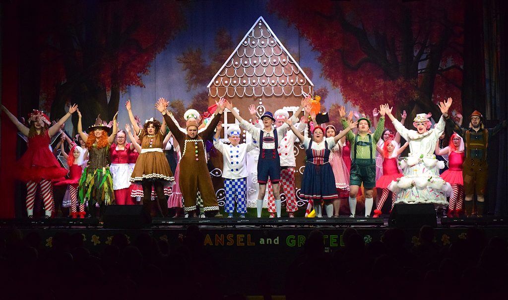 front of the stage, all with their arms raised in a final bow. The performers are dressed in colourful fairy-tale costumes including gingerbread characters, bakers, villagers, woodland characters and candy-themed outfits. Behind them is a large gingerbread house set against an autumn woodland backdrop. Stage lighting highlights the cast, with the audience visible in shadow at the bottom of the image.