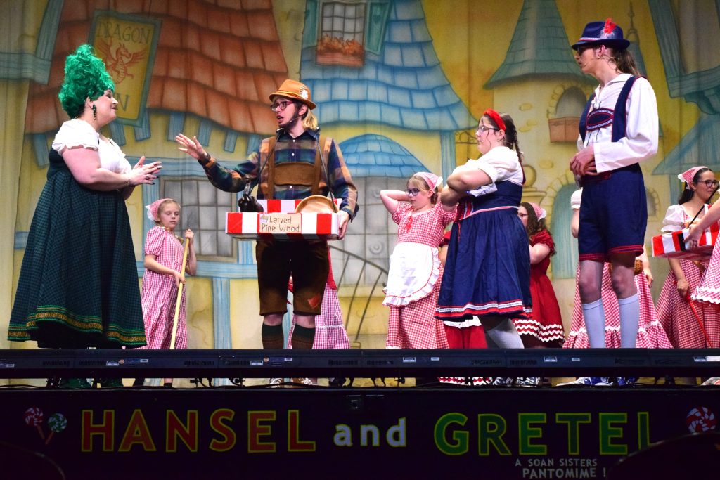 A stage scene from a Hansel and Gretel pantomime showing a group of performers in traditional fairy-tale costumes. At centre, a man dressed as a woodcutter or trader gestures while holding a red-and-white striped box labelled “Carved Pine Wood,” speaking to a woman in a green dress with a bright green wig. Around them, children and adults dressed as villagers and bakers in red-and-white gingham costumes look on, some with arms folded. The backdrop shows colourful illustrated village houses, and the stage banner below reads “Hansel and Gretel – a Soan Sisters pantomime.”