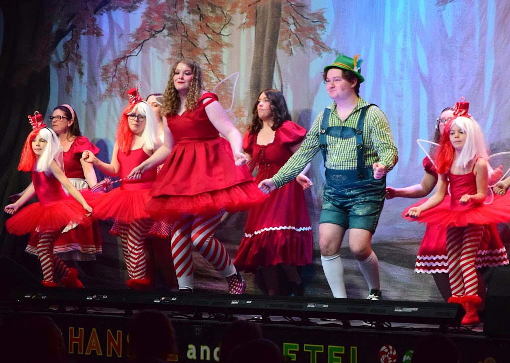 A lively dance scene from a Hansel and Gretel pantomime shows a group of performers moving in unison across a forest-themed stage. Several dancers wear bright red dresses with tulle skirts, striped red-and-white tights, and fairy wings, while others wear matching red costumes with festive hair accessories. One performer at centre-right wears green lederhosen with a checked shirt and a green hat, stepping in time with the group. The cast smile and concentrate on the choreography, with the colourful woodland backdrop and stage lighting enhancing the energetic, playful atmosphere.