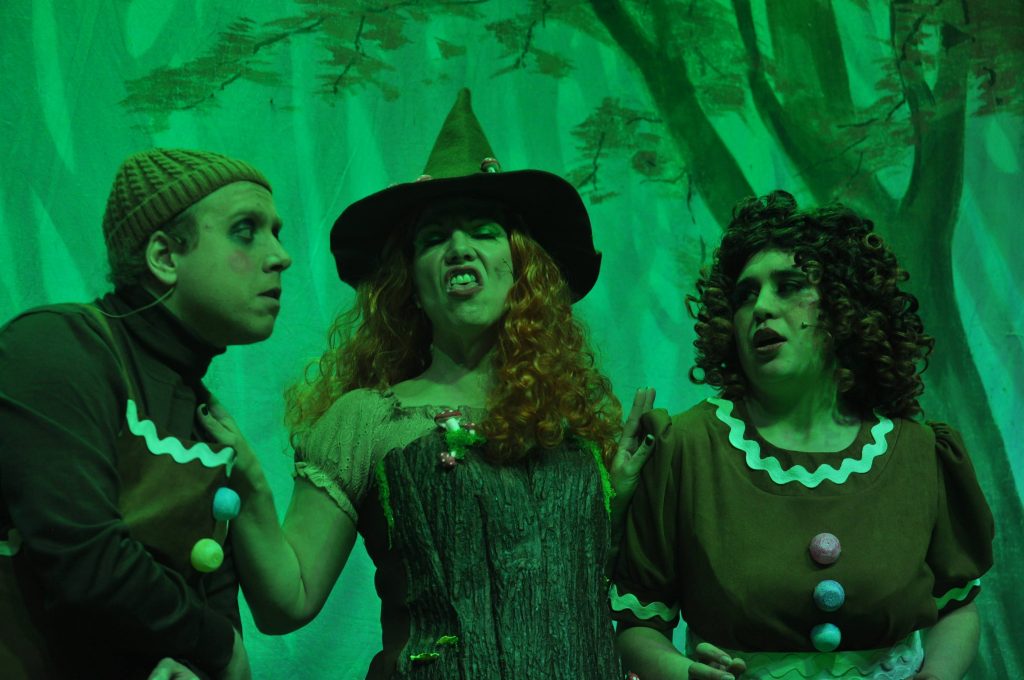 A close-up stage scene from a Hansel and Gretel pantomime lit in dramatic green lighting. Three performers stand together against a painted forest backdrop: at centre, a woman dressed as the Witch wears a pointed hat and a bark-textured dress, pulling a theatrical grimace as she gestures mid-song or dialogue. On either side of her, two performers dressed as gingerbread characters in brown costumes with icing-style trim look towards her with concerned and wary expressions, suggesting a tense but comedic moment in the scene.