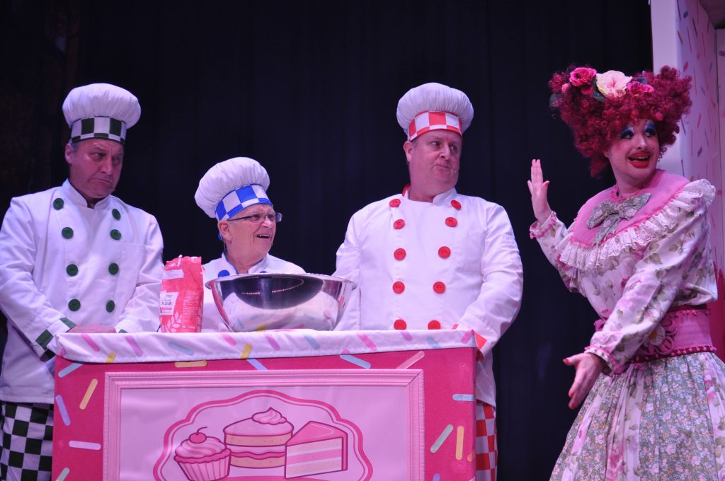 A brightly lit stage scene from a Hansel and Gretel pantomime shows three performers dressed as bakers behind a pink, sprinkle-decorated baking counter with a large mixing bowl and a bag of flour. The bakers wear white chef’s outfits with tall hats trimmed in green, blue, and red, and react with amused and sceptical expressions. To the right, a performer in an elaborate floral dress with a wide pink belt and a large curly red wig adorned with flowers gestures theatrically towards them, mid-performance, creating a playful and comedic moment.