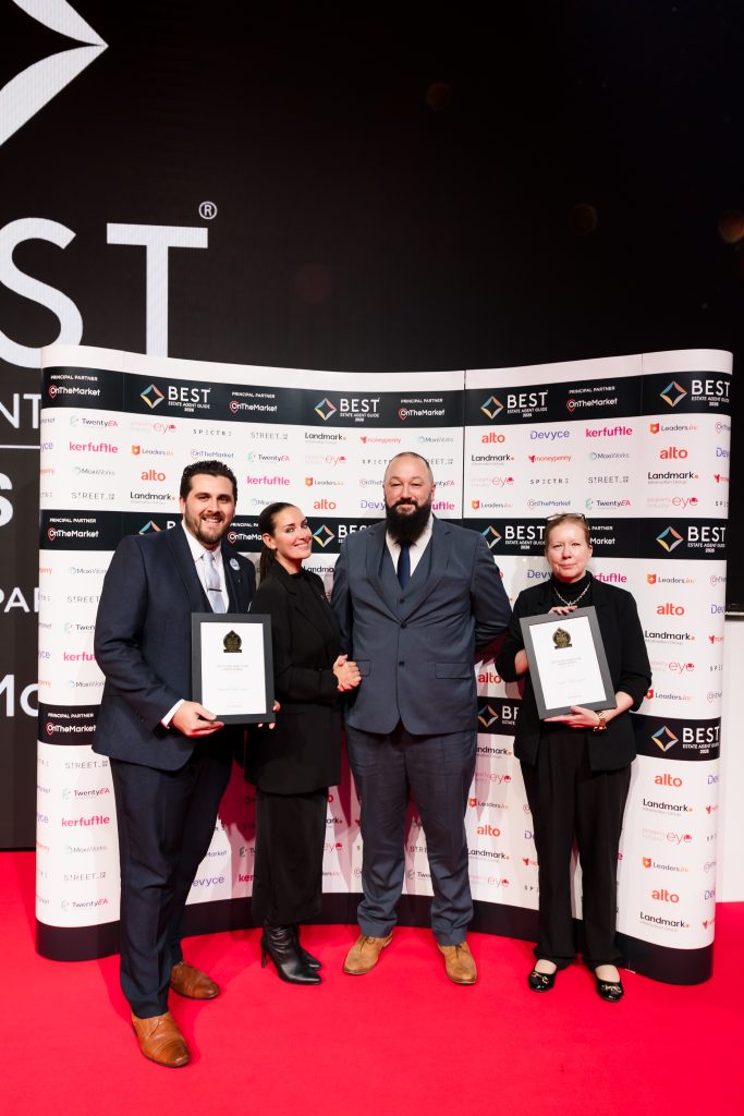 Four people stand on a red carpet at an awards event in front of a branded “BEST Estate Agent Guide” step-and-repeat backdrop featuring multiple sponsor logos. Two of them hold framed award certificates. All are dressed in formal attire: two men in suits (one with a beard), and two women in black outfits. They are smiling and posing for a presentation photo at what appears to be an estate agency industry awards ceremony.