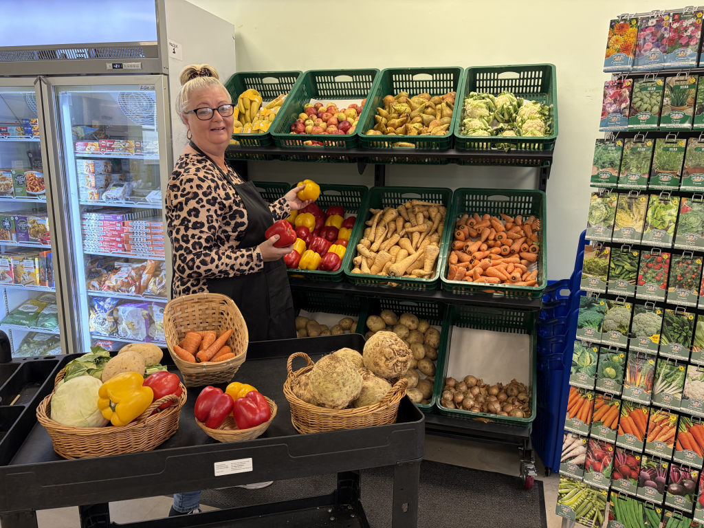 A woman wearing glasses, a leopard-print top and a black apron stands inside a small shop beside neatly stacked green crates of fresh produce. She holds a red and a yellow pepper while standing in front of shelves filled with apples, pears, bananas, cabbages, parsnips, carrots, potatoes and onions. In front of her, baskets on a display table contain carrots, peppers, celeriac and cabbage. A glass-door freezer is visible to the left, and a tall rack of vegetable and flower seed packets stands to the right.