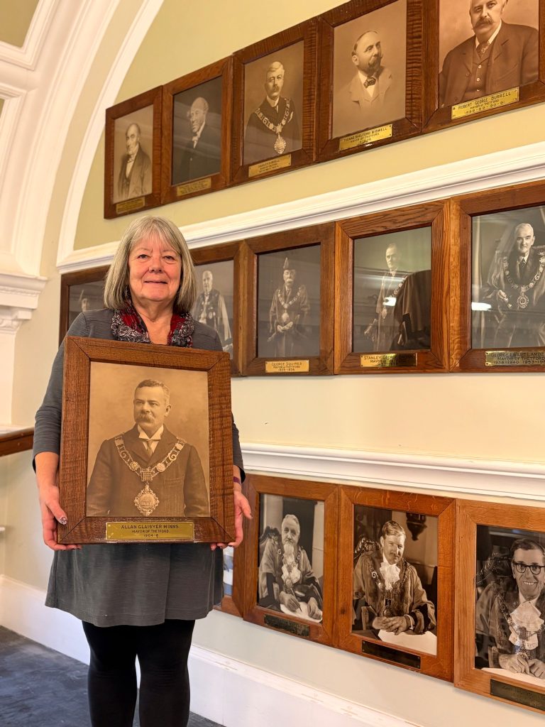A woman with shoulder-length grey hair stands indoors against a cream wall lined with framed sepia and black-and-white portraits of former mayors in wooden frames. She is holding a large framed sepia portrait of Allan Glaisyer Minns, Mayor of Thetford (1904–06), wearing mayoral chains, with a brass nameplate beneath the image. The surrounding wall displays multiple historic mayoral portraits arranged in neat rows, each with engraved plaques, in what appears to be a civic building or council chamber.