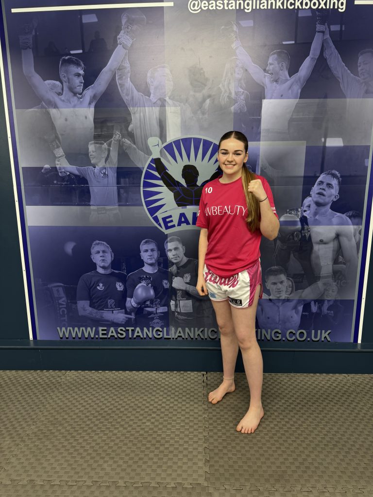 A young female kickboxer stands barefoot on gym mats in front of a large East Anglian Kickboxing wall display featuring action photos of fighters and the club logo. She is wearing a bright pink T-shirt and pink-and-white kickboxing shorts, smiling and posing with one fist raised. The website address www.eastangliankickboxing.co.uk is visible along the bottom of the backdrop.