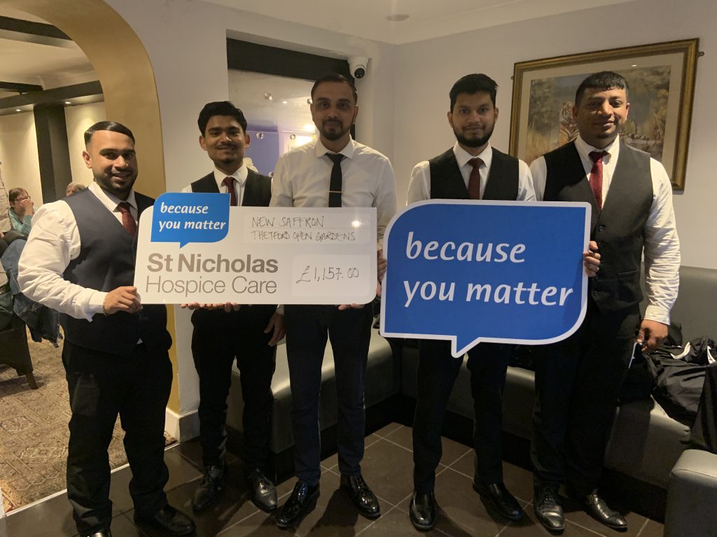 Five men dressed in white shirts, black waistcoats and ties stand inside a restaurant holding a large cheque for St Nicholas Hospice Care. The cheque reads “New Saffron Thetford Open Gardens” and shows a donation of £1,157.00. Two of the men hold blue speech-bubble signs reading “because you matter,” the hospice’s slogan.