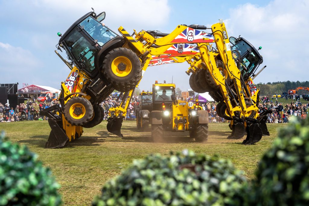 Two bright yellow JCB diggers perform a stunt at an outdoor show, balancing on their front buckets with their rear wheels lifted high off the ground, facing each other like mirrored arches. A third JCB vehicle drives between them. A large crowd watches from behind barriers in the background under a blue sky, suggesting a live arena demonstration at a country fair.