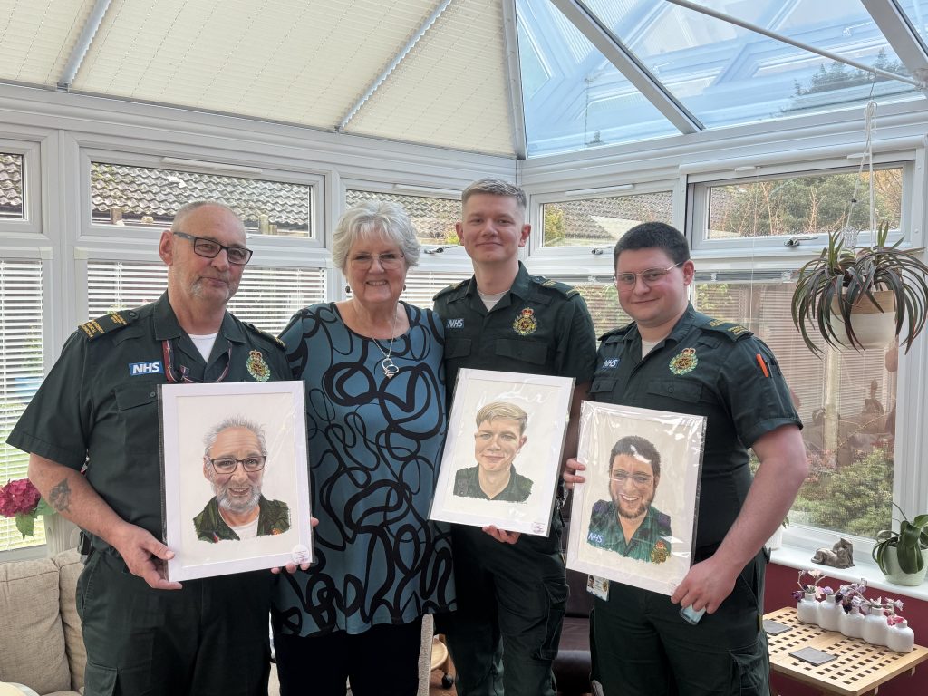 Four people standing together in a bright conservatory with large windows and blinds. Three NHS uniformed staff members in green paramedic-style shirts stand either side of an older woman in a patterned blue and black top. Each of the uniformed staff holds a framed illustrated portrait of themselves. Potted plants and small decorative items sit on a table and windowsill behind them, and rooftops and trees are visible through the windows.
