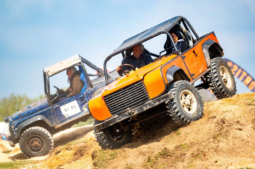 Two off-road vehicles tackle a sandy obstacle course during a countryside event. In the foreground, an orange Land Rover-style 4x4 climbs a steep dirt mound at an angle, its large tyres gripping the loose ground as the driver concentrates at the wheel and a passenger sits beside them. Behind it, a blue 4x4 follows over another mound. The scene appears to be part of a live off-roading demonstration under a clear blue sky.