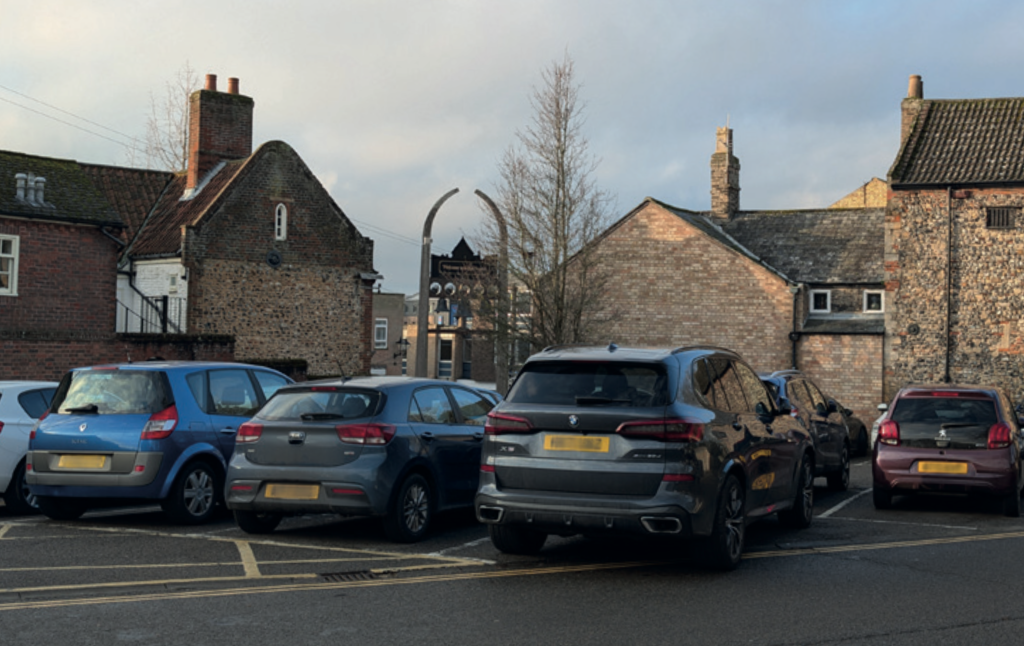 A photograph showing a small town car park with several parked cars lined up in marked bays, including blue, grey, black and red vehicles. The car park is bordered by older brick and flint buildings with pitched roofs and chimneys, typical of historic town architecture. In the background, a narrow street leads between the buildings, with a leafless tree visible under a pale winter sky.