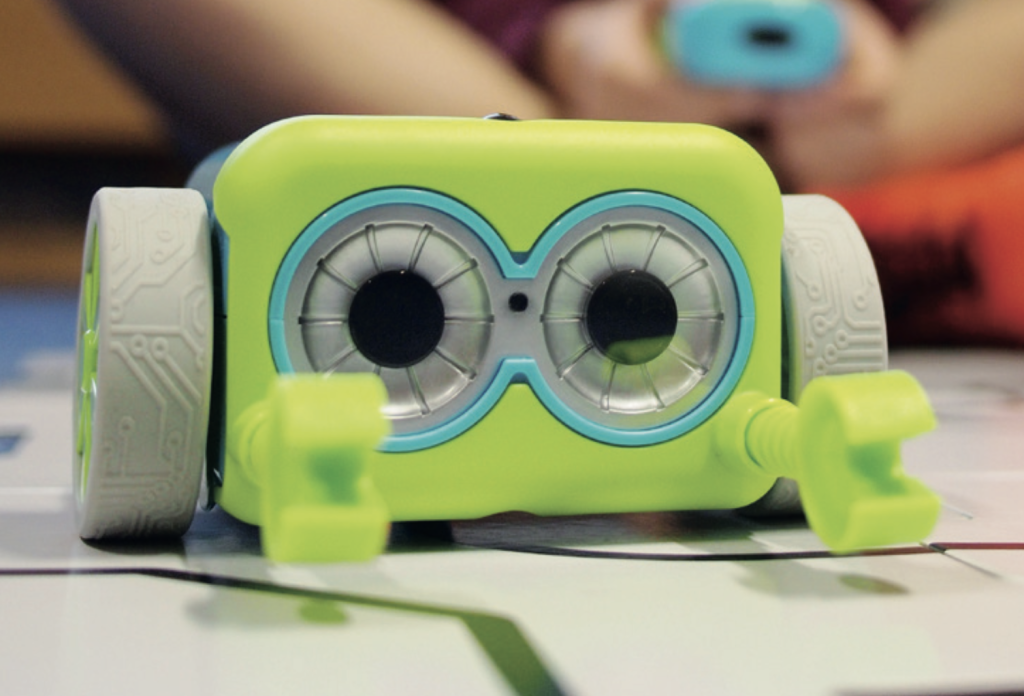 Close-up of a small lime green educational robot with two large circular “eyes” and grey wheels on either side, sitting on a tabletop with a printed track. A child’s hands are blurred in the background, suggesting the robot is being used for a coding or learning activity.