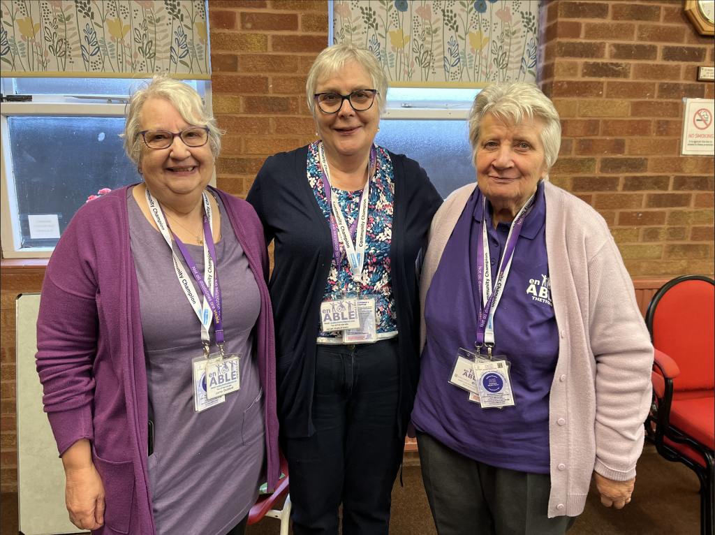 Three older women stand side by side in a brick-walled community hall, smiling at the camera. All three wear lanyards with name badges, and two are dressed in purple tops featuring the enABLE Thetford logo. Behind them are patterned window blinds, a “No Smoking” sign on the wall and red chairs, suggesting a welcoming community group setting.