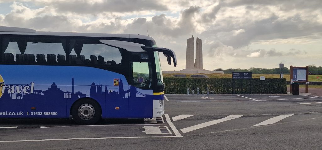 A blue coach parked in a roadside car park with a tall stone war memorial visible in the background under a cloudy sky.