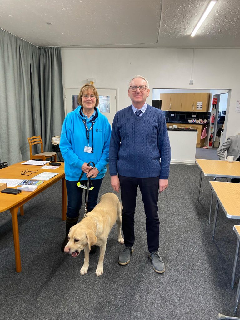 Two people stand side by side in a community hall, smiling at the camera, with a yellow Labrador guide dog on a lead in front of them. The woman wears a blue Guide Dogs hoodie and holds the dog’s lead, while the man stands beside her in a jumper and shirt. Tables with leaflets and chairs are visible around the room.