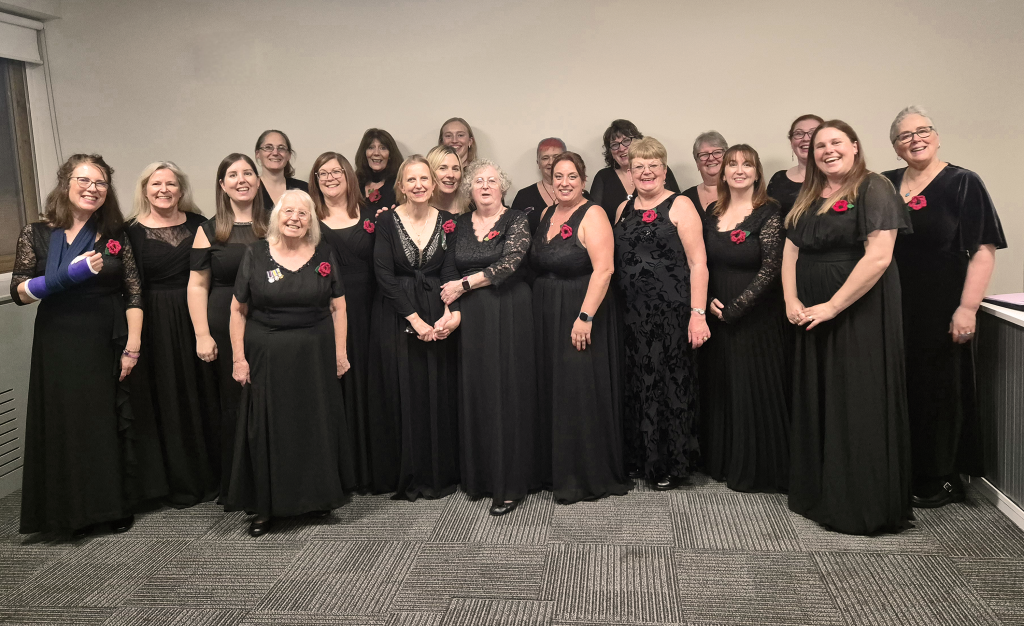 A large group of women stand together in a line indoors, dressed in long black concert dresses with red flower pins, smiling for a photo after a performance. The group appears to be a choir or singing group gathered against a plain wall.