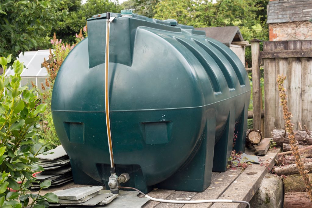 A large green domestic oil storage tank with a fuel gauge and pipe attached, positioned in a garden beside plants, fencing and stacked materials.