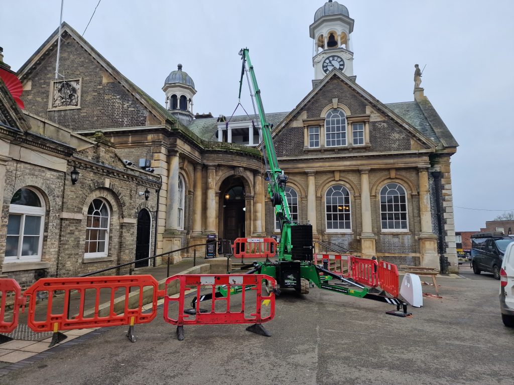 Front view of a historic civic building with a clock tower and classical stone façade. A green crane is positioned in the courtyard, lifting a dormer window structure onto the roof. Red safety barriers surround the work area, with a van parked to the right, indicating ongoing maintenance or restoration work.