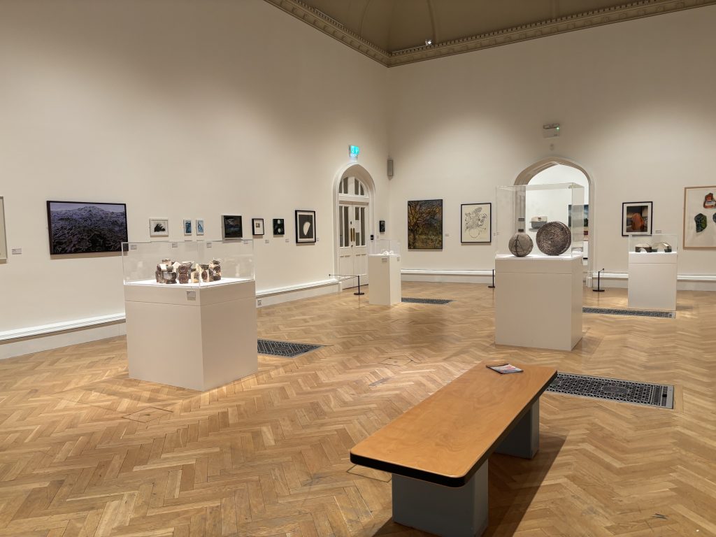 Interior of an art gallery with high ceilings and a polished wooden parquet floor. Various artworks are displayed around the room, including framed pieces on the walls and sculptures in glass display cases on white plinths. A wooden bench sits in the foreground, and doorways and archways are visible along the back wall.