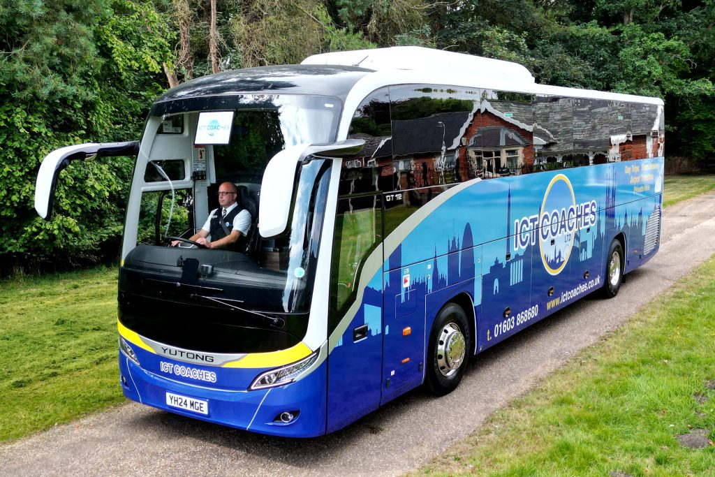 A blue ICT Coaches tour bus drives along a narrow rural road bordered by grass and trees, with the driver visible through the front windscreen.