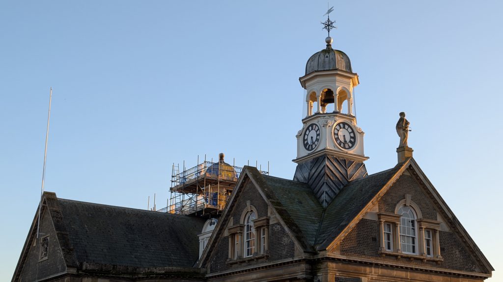Historic building with a clock tower, photographed in warm evening light against a clear sky. The tower features a domed top with a weather vane and visible clock faces on two sides. A statue stands on the roofline to the right, while scaffolding is visible behind part of the building, indicating maintenance work.