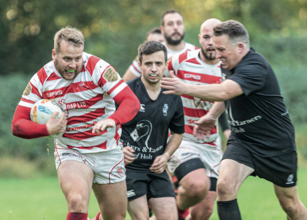 A rugby player in a red-and-white hooped jersey runs forward with the ball during a match, while several opposing players in black kits chase and attempt to tackle him on a grassy pitch.