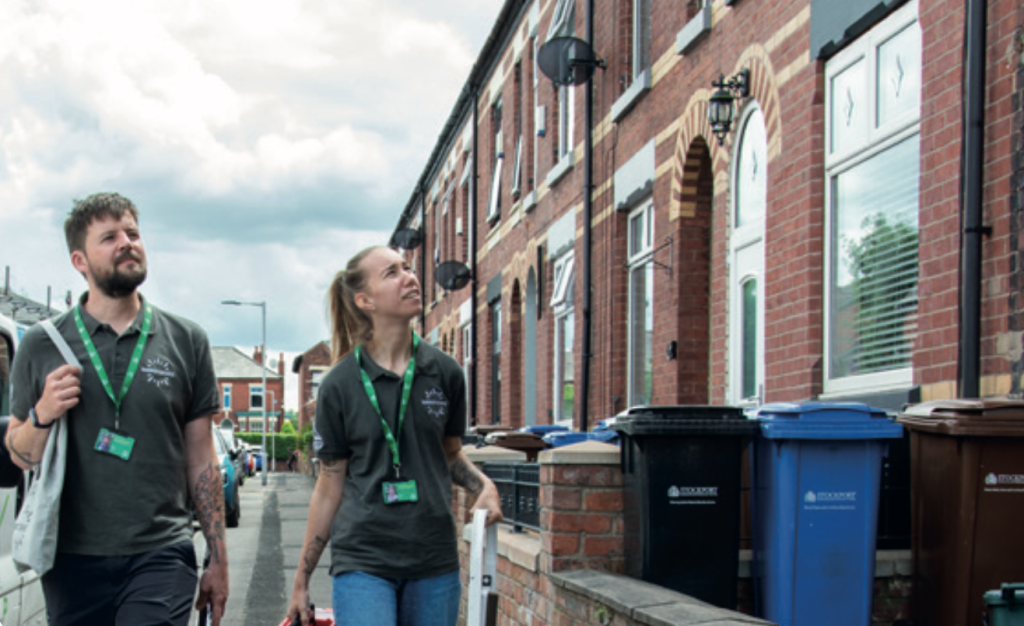 Two outreach workers wearing green lanyards walk along a residential street of red-brick terraced houses, looking up at the properties. Wheelie bins line the pavement outside the homes, and the sky is overcast.
