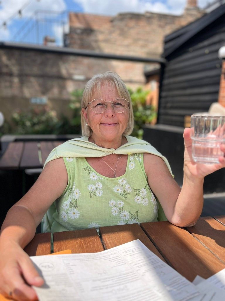 An older woman wearing glasses and a green floral top sits at an outdoor table in the sunshine, smiling and holding up a glass of water. Papers are spread out on the table in front of her, with a garden and buildings softly blurred in the background.