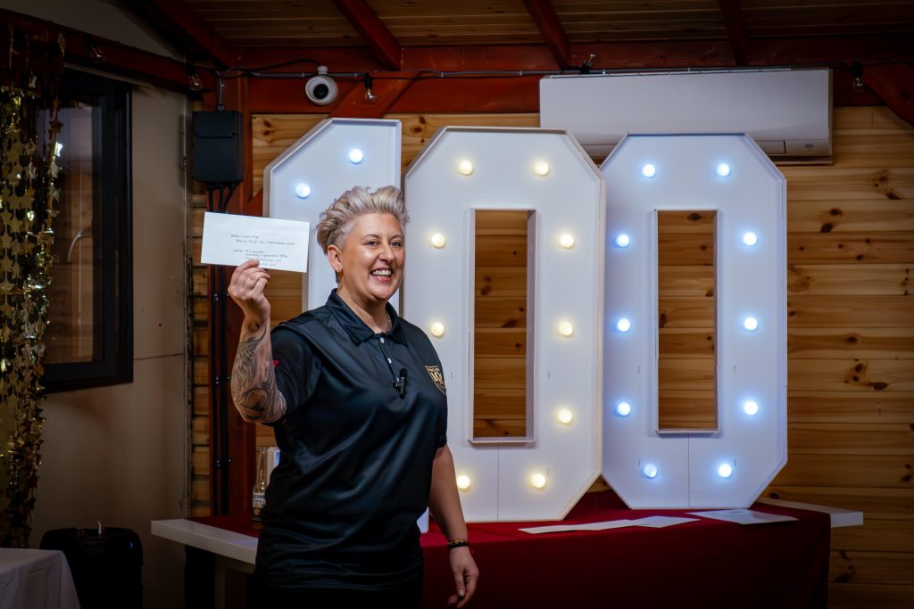 A smiling woman stands indoors in front of large illuminated “100” numbers, each outlined with small round bulbs. She is holding up a white envelope in her right hand, angled slightly towards the camera, as if presenting it. The handwritten text on the envelope is partially visible but not fully legible.She has short, light blonde hair styled upwards and is wearing a dark polo shirt with a small embroidered logo on the left chest. A microphone is clipped to her shirt, suggesting she may be speaking or presenting. A tattoo is visible on her right forearm.Behind her, the large “100” display sits on a table covered with a red cloth. The setting appears to be a wooden interior space, with timber walls and exposed beams. Above the numbers is a wall-mounted air conditioning unit, and to the left there is a door with a window. Decorative string lights and a small security camera are also visible along the beam.The overall scene suggests a celebratory or fundraising moment, with the “100” display acting as a focal point, marking a campaign target.