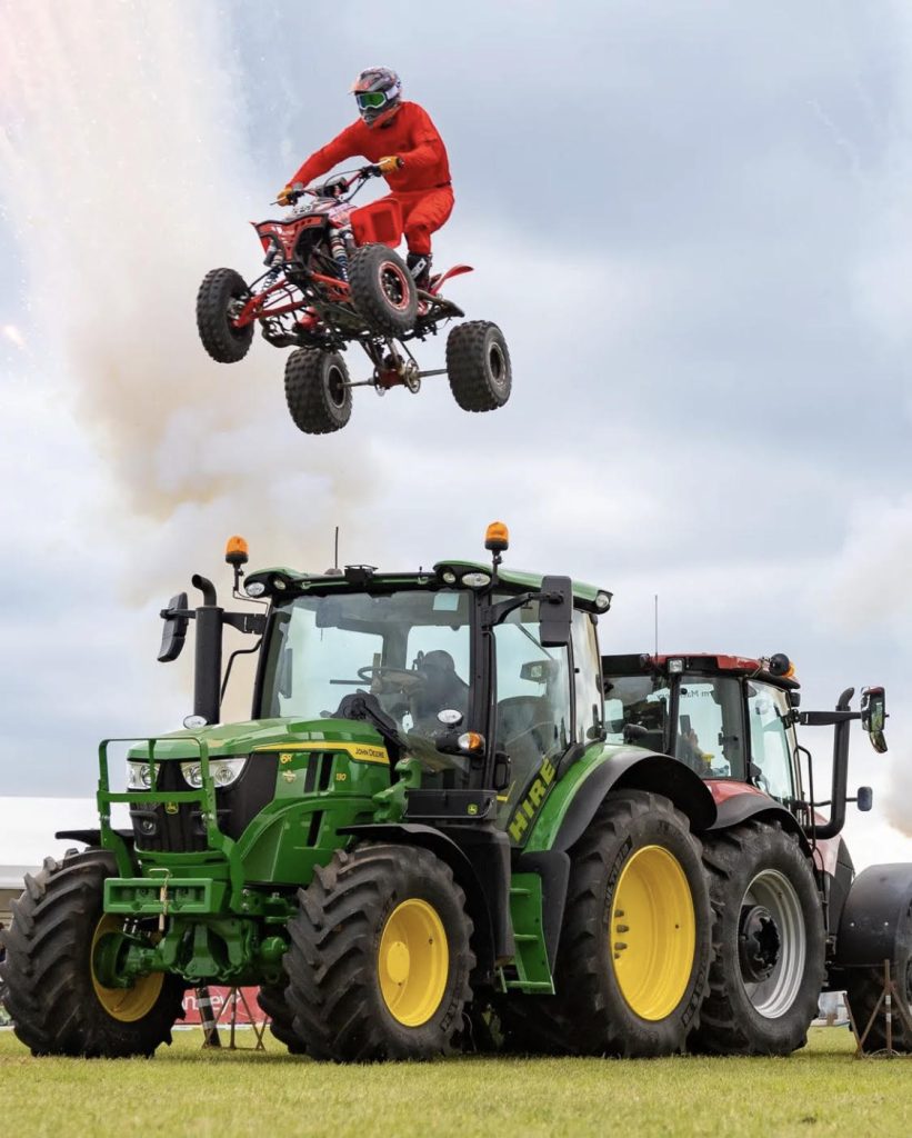 A quad bike rider in red protective gear is captured mid-jump high in the air above a green tractor during a stunt display, with smoke or dust in the background.