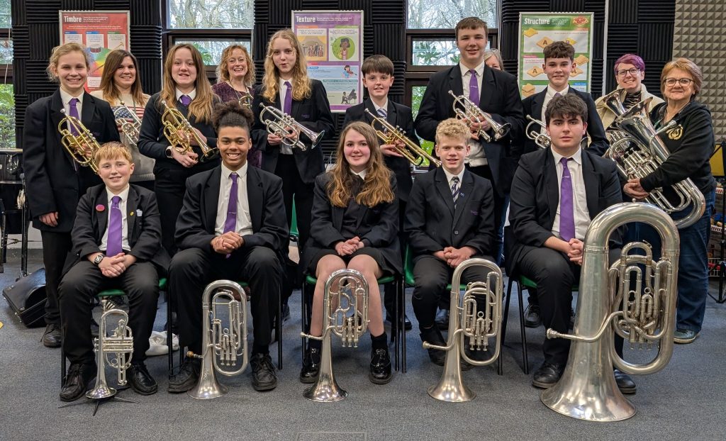 A group of school students and adults pose indoors with brass instruments in what appears to be a music classroom or rehearsal space.In the front row, five students sit on chairs holding larger brass instruments such as baritones and euphoniums, with one very large tuba positioned on the far right. They are wearing school uniforms consisting of black blazers, white shirts, and purple ties. The students are smiling or looking towards the camera, seated with instruments resting on the floor in front of them.Behind them, a second row of students stands holding smaller brass instruments, including cornets or trumpets. They are also in matching school uniform. Two adult women stand among them—one in the centre smiling broadly, and another on the far right holding a large brass instrument and wearing glasses.The background shows classroom walls with acoustic foam panels and educational posters titled “Timbre”, “Texture”, and “Structure”. Windows behind the group reveal trees outside, indicating daytime.The overall scene suggests a school band or music group posing for a photo, likely celebrating participation or achievement in a brass ensemble.