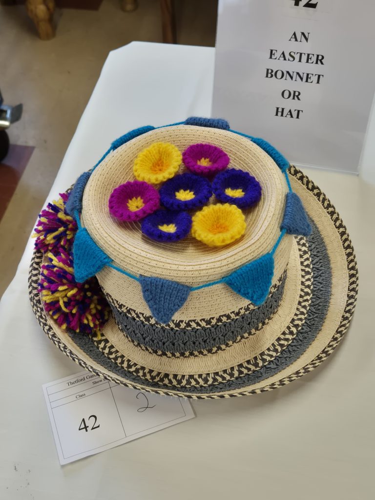 A wide-brimmed straw hat decorated with colourful knitted flowers and bunting is displayed on a table, with a sign behind reading “An Easter Bonnet or Hat” and a competition entry card marked “42” in front.