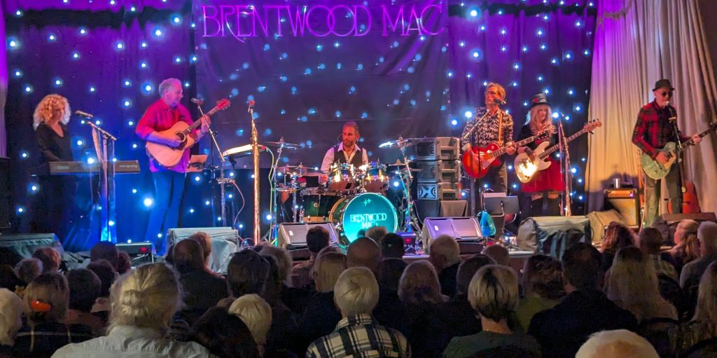 A live band performs on stage in front of a seated audience, with a backdrop reading “BRENTWOOD MAC” and a curtain of small blue stage lights. The group includes a keyboard player, acoustic guitarist, drummer, and multiple electric guitarists and singers. Stage lighting casts purple and blue tones across the performers, while the crowd fills the foreground watching the show.